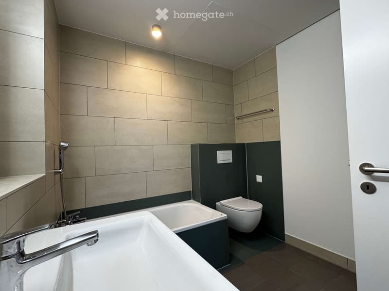 Modern bathroom with beige tiled walls, a large bathtub, a toilet, and a showerhead. Features a sleek, contemporary design with a dark accent wall and a built-in shelf.