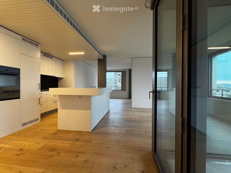 Modern kitchen with white cabinets, a central island, and an oven, connected to a living area with wooden flooring and large windows offering a city view.