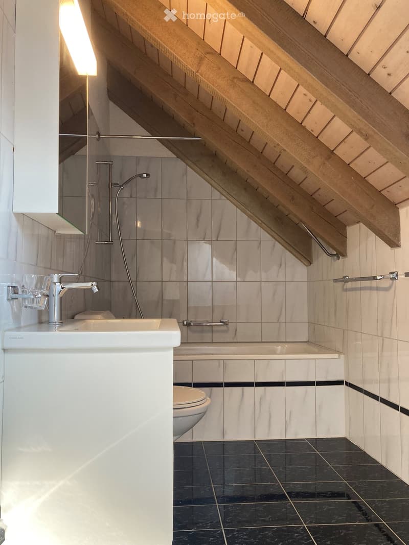Modern bathroom with a white sink, chrome faucet, toilet, bathtub, and handrail. The walls are tiled in light grey with a dark grey accent border, and the floor is dark grey tiles. The ceiling has exposed wooden beams.