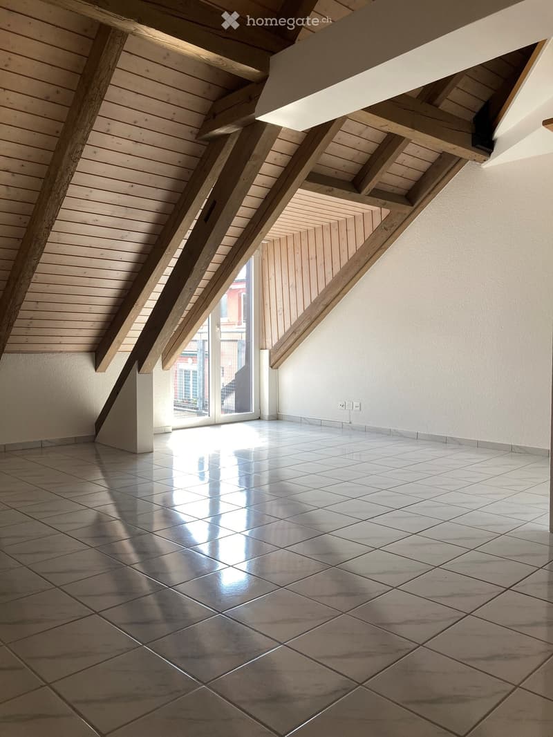 Empty room with polished tile flooring, exposed wooden beams, and white walls. Natural light streams in from a large window or door at the back.