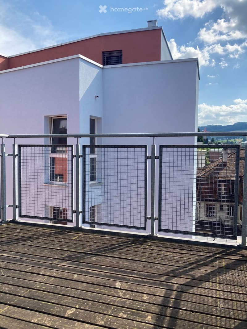 Wooden deck with metal railing, overlooking a modern building with white and red walls and multiple windows.