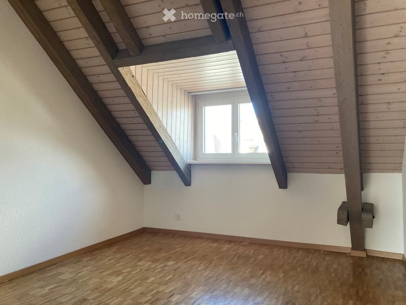 Empty attic room with wooden flooring, white walls, and exposed wooden beams. Features a single window with white frame and blinds.
