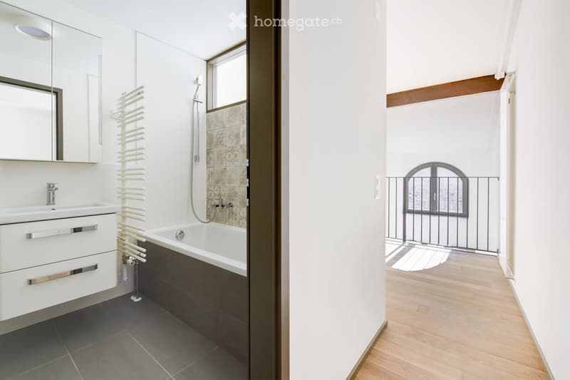 Modern bathroom with a white sink, bathtub, mirror, and radiator. The space features dark tiled flooring and a wooden-floored hallway leading to a balcony.