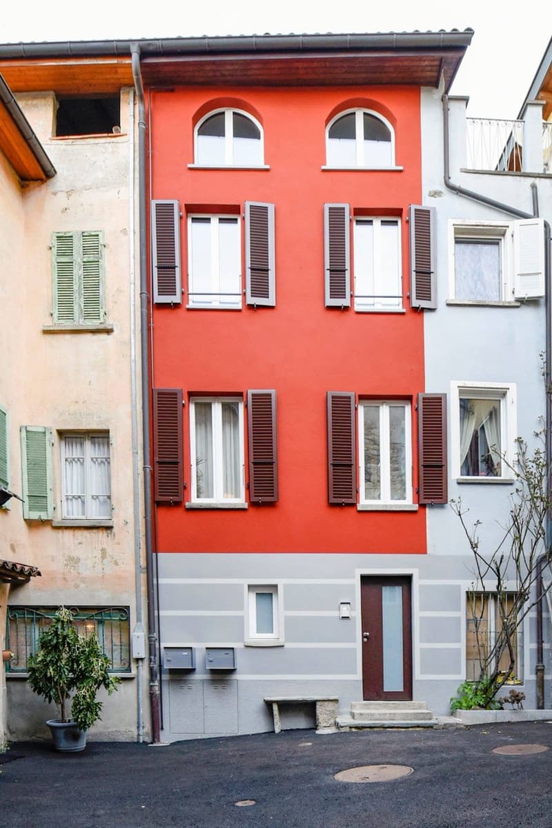 A three-story residential building with a red upper facade, grey lower facade, multiple windows with shutters, and a central entrance door.