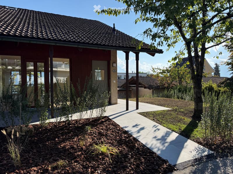 Modern house exterior with a tiled roof, wooden facade, and a landscaped garden area featuring mulch beds, small plants, and a paved walkway.