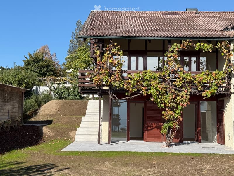 Two-story house with a tiled roof, wooden balcony, and a small tree in the front yard. The house has large windows and a raised ground floor with steps leading up to the entrance.