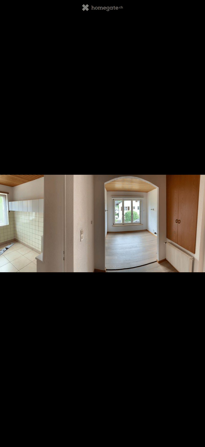 Empty hallway with wooden flooring, white walls, and a view into an adjacent room with large windows and a radiator.