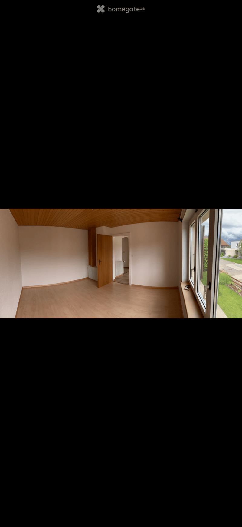 Empty room with wooden flooring, white walls, and large windows offering a view of a green garden. The room features a wooden door leading to another area and a slanted wooden ceiling.