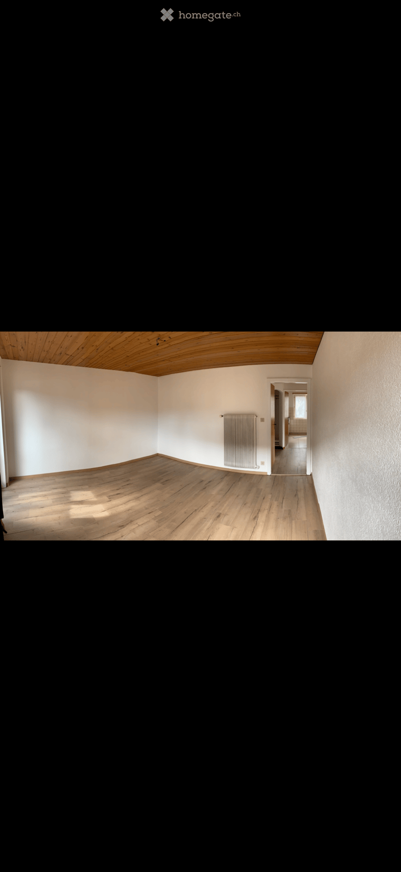 Empty room with wooden flooring, white walls, and a radiator. The room has a wooden slatted ceiling and a doorway leading to another room.
