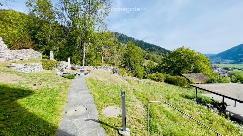 A scenic outdoor area with a stone pathway, grassy lawn, benches, and a view of a valley and mountains.