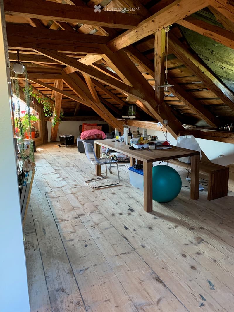 Loft-style room with exposed wooden beams and wooden flooring, featuring a long wooden table with chairs, a large exercise ball, and various items on the table including bottles and a lamp.
