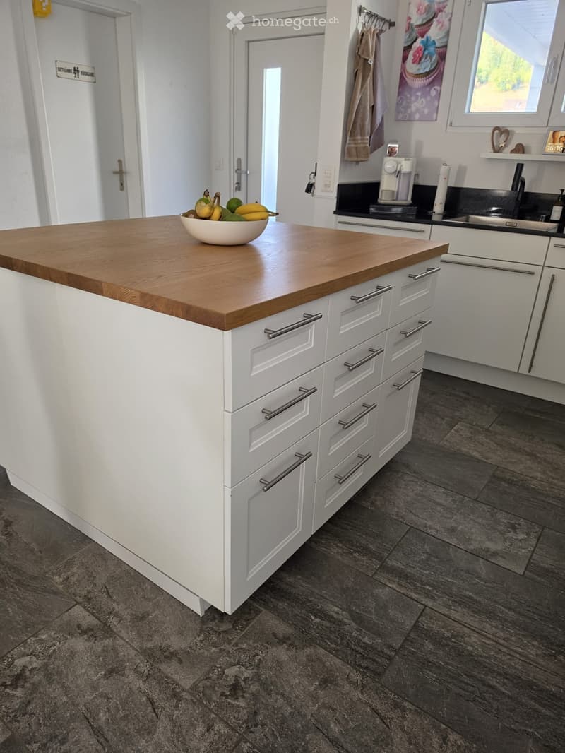 Modern kitchen with a white island featuring a wooden countertop, a bowl of fruit, and various appliances including a coffee maker and sink.