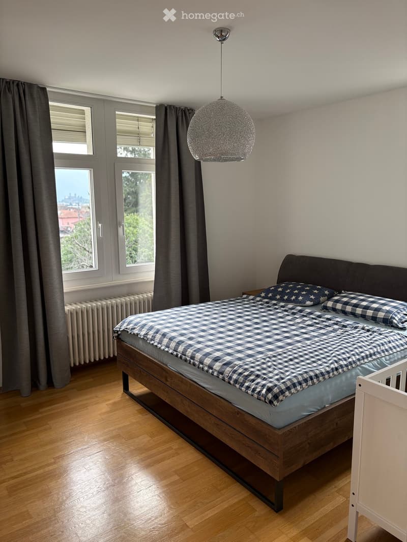 A well-lit bedroom with wooden flooring, a large bed with blue and white checkered bedding, a nightstand, and a modern pendant light fixture.