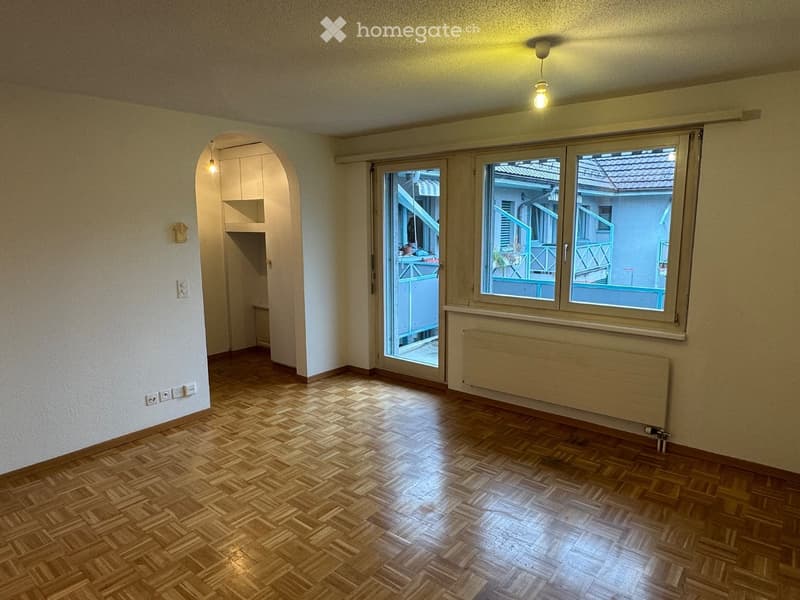 Empty room with wooden parquet flooring, white walls, large window with view to balcony, radiator, and arched doorway leading to another space.