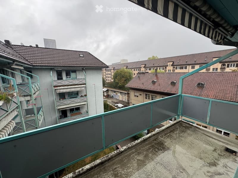 View from a rooftop terrace overlooking a multi-story residential building with balconies, striped awnings, and surrounding structures.