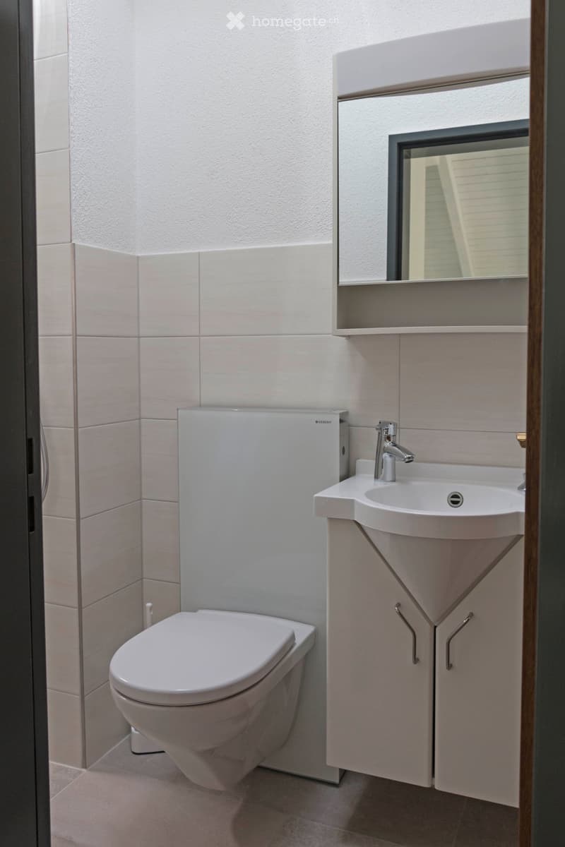 Modern bathroom with a toilet, sink, and mirror. The sink has a sleek faucet and is mounted on a white cabinet. The walls are tiled in light beige, and the floor is also tiled.