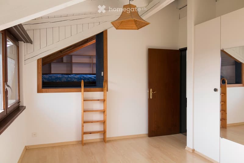 A small, cozy room with light wooden flooring and white walls. Features a lofted bed with a ladder, a hanging pendant light, and a wooden door leading to another space.