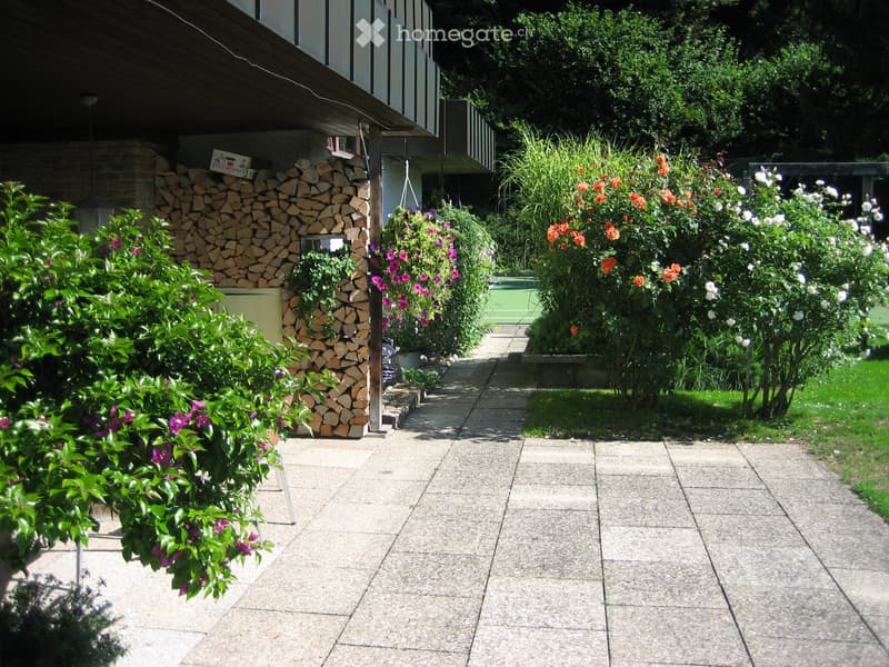 Garden path lined with colorful flowers and shrubs, with a woodpile and a building in the background.