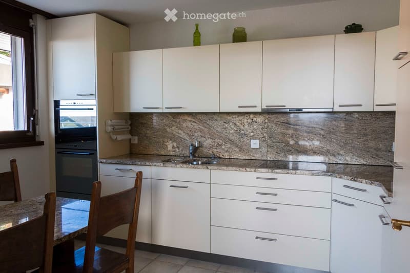 Modern kitchen with white cabinets, granite countertops, and a sink with a faucet. There is a wooden dining table with chairs in the foreground.