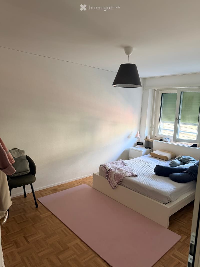 A modern bedroom with a white bed, wooden floor, pink rug, and a hanging lamp. A chair with a handbag is placed near the bed, and a small nightstand with a lamp is visible.