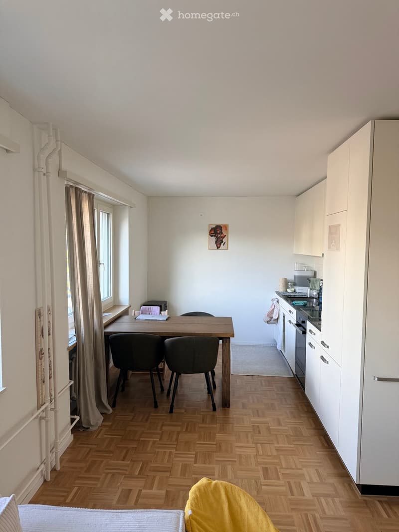A modern, open-plan room with a wooden floor, a dining table with chairs, a kitchen area with cabinets, a refrigerator, and a sink, and a bed with a yellow blanket in the foreground.