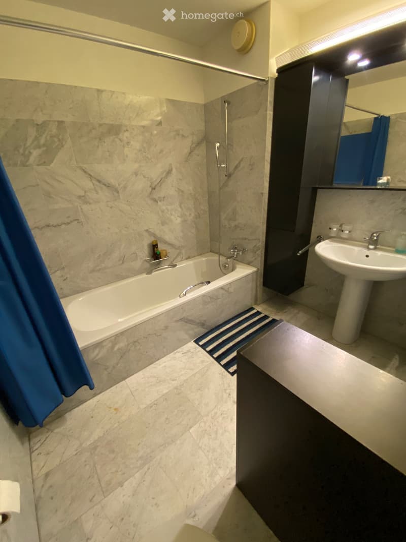 Modern bathroom with marble walls and floor, bathtub, sink, mirror, and dark cabinetry.