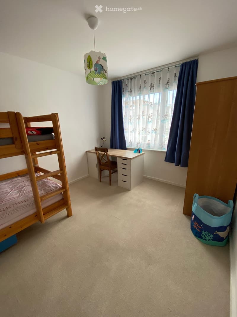 A small bedroom with a wooden bunk bed, a desk with a chair, and a window with curtains.