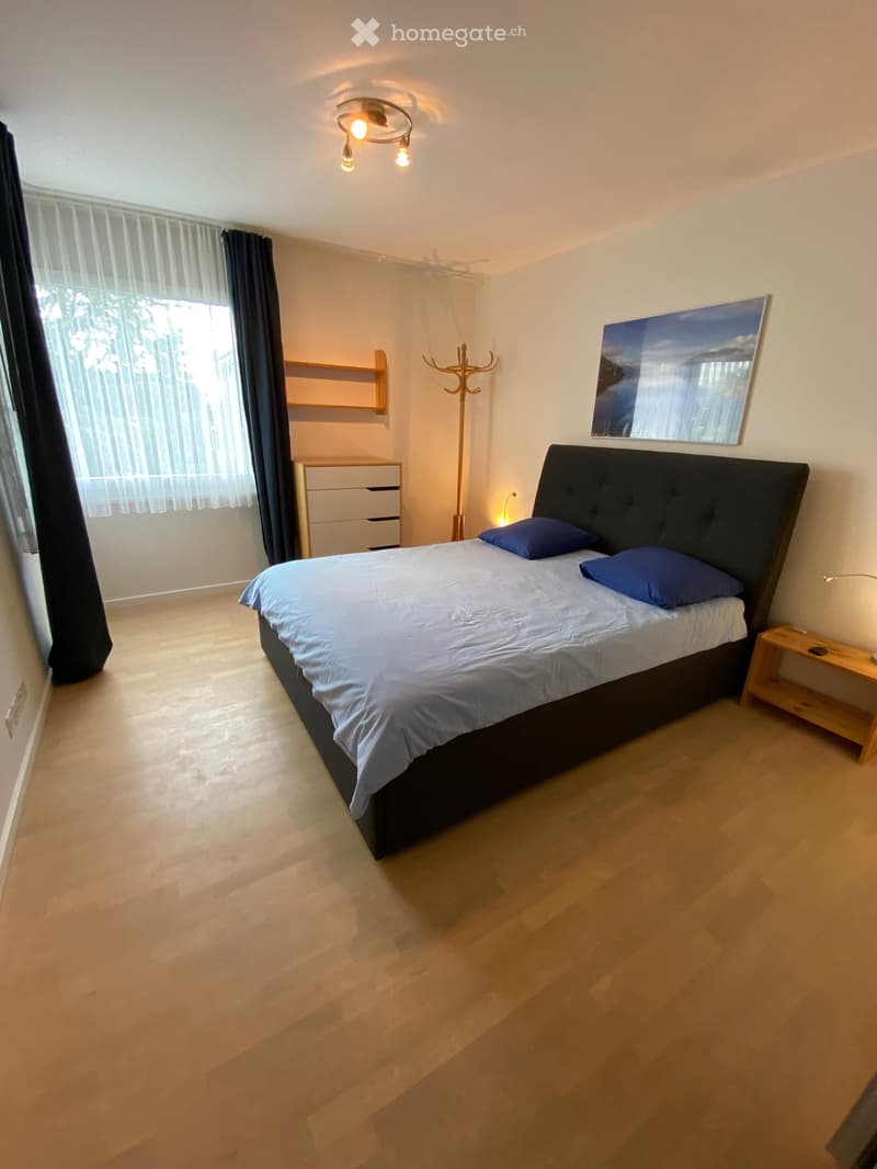 Modern bedroom with a large bed, nightstand, lamp, and large window with curtains. Features a coat rack, shelf, and picture frame on the wall.
