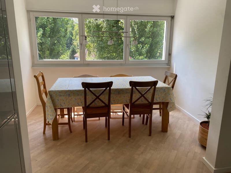 Bright dining room with wooden flooring, large windows, a dining table with a floral tablecloth, and six wooden chairs.