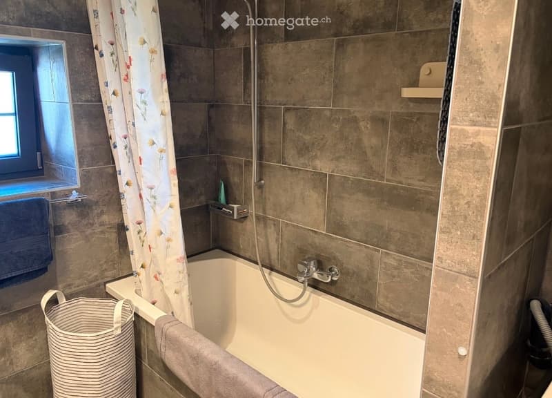 Modern bathroom with a bathtub, shower curtain, towels, a basket, and a window providing natural light.