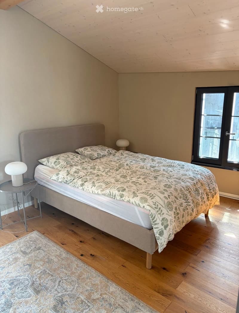 Cozy bedroom with wooden flooring, a large bed with a patterned duvet, a nightstand with a lamp, and a soft rug on the floor.