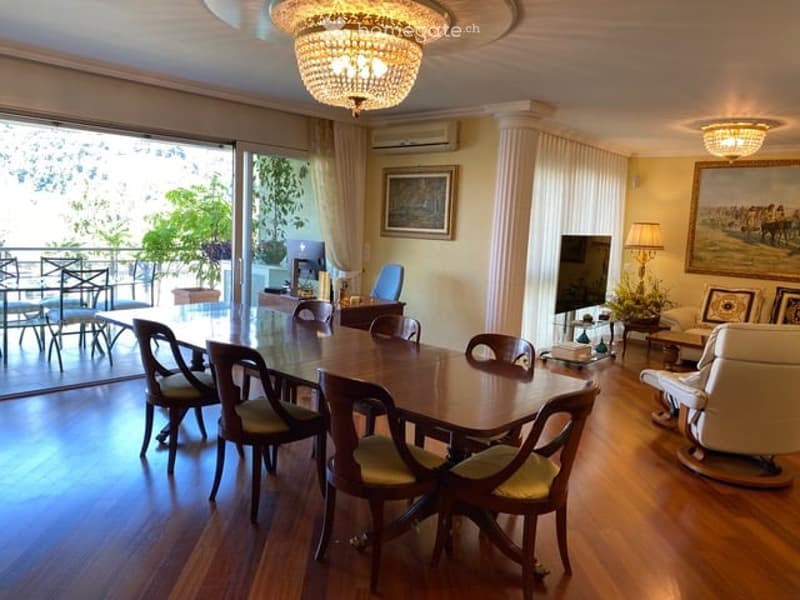 Spacious dining and living area with wooden flooring, large glass doors leading to a balcony, a dining table with chairs, a sofa, a television, and decorative lamps.