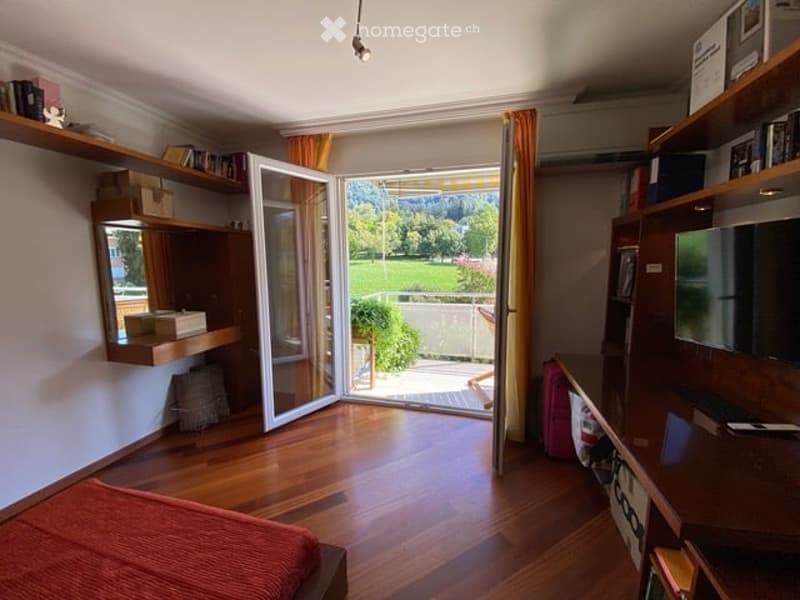 Bright bedroom with wooden flooring, large sliding glass doors opening to a view of greenery, a bed with an orange blanket, a desk with a laptop, shelves with books and decorative items, and a mirror on the wall.