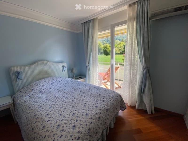 Cozy bedroom with light blue walls, wooden flooring, a neatly made bed with a floral duvet, a nightstand with a lamp, and large windows with curtains offering a view of a green outdoor area.
