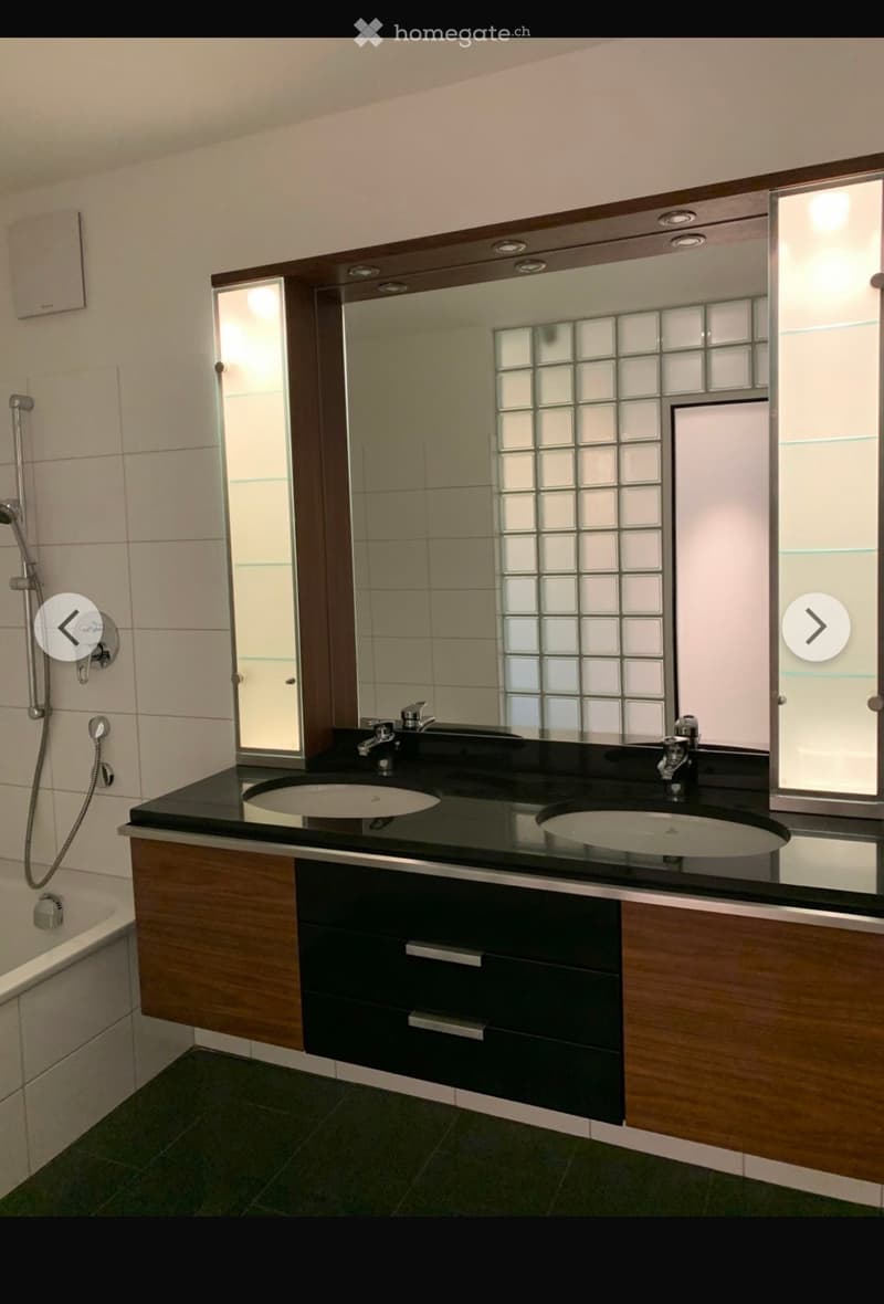 Modern bathroom with double sink vanity, black countertop, wooden cabinets, large mirror, and glass block window.