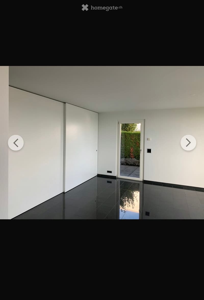 Empty room with white walls, black tiled floor, and a large sliding glass door leading to a garden area.