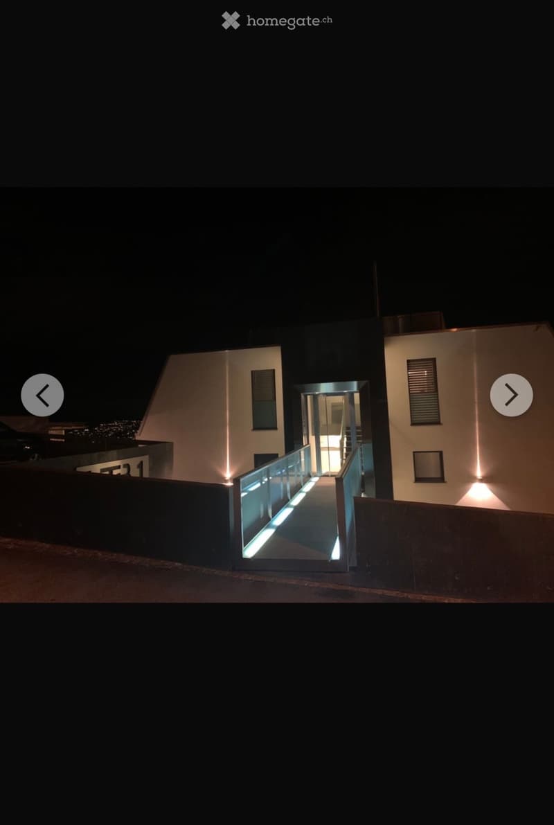 Modern two-story building with illuminated entrance and sleek design, featuring a glass walkway leading to the entrance.