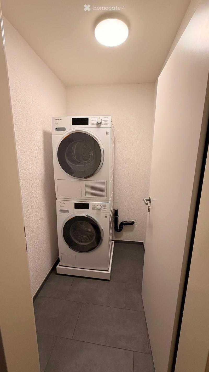 Compact bathroom with a stacked washing machine and dryer unit, white walls, grey tiled floor, and a round ceiling light.