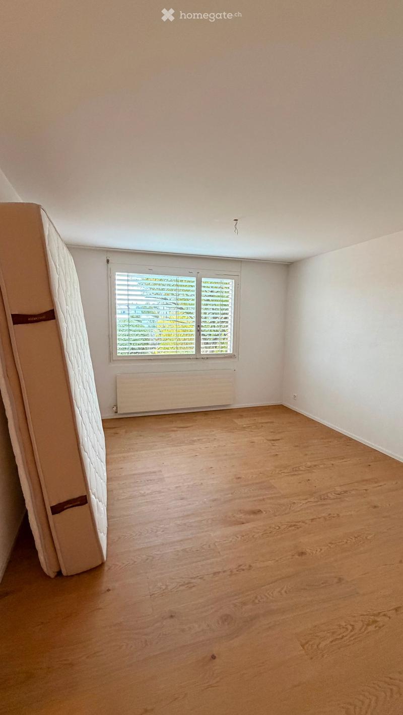 Empty room with wooden flooring, white walls, a window with blinds, and a radiator under the window. A mattress is partially visible on the left side.