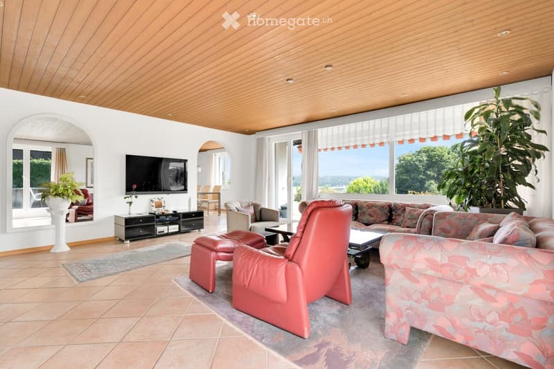 Bright living room with large windows and a scenic view, featuring a red leather chair, floral-patterned sofa, coffee table, side table, and television.
