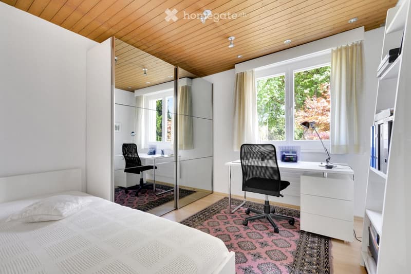 Modern bedroom with a bed, desk, chair, and shelves. Features large windows with curtains, a wooden ceiling, and a patterned rug.
