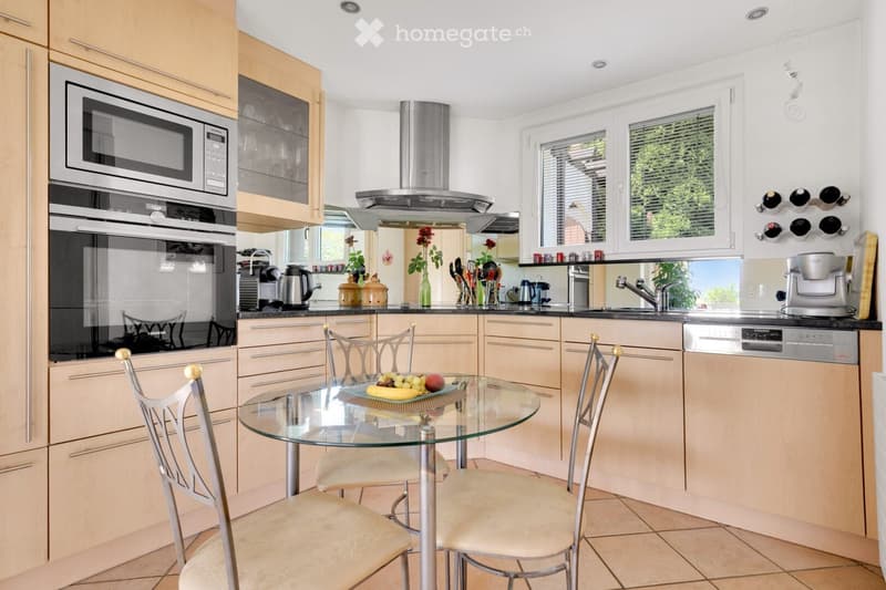 Modern kitchen with light wood cabinets, stainless steel appliances, a glass dining table with four chairs, and a view of greenery through the windows.