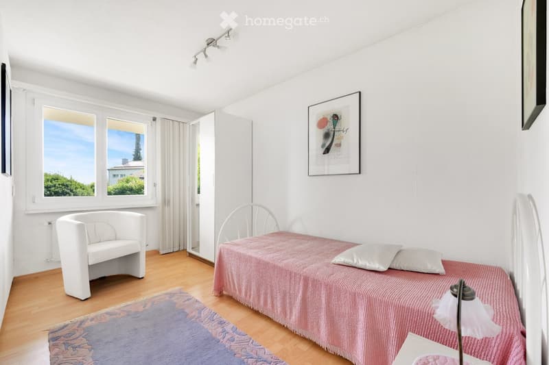 A small, modern bedroom with a single bed, a white armchair, a bedside lamp, and a picture frame on the wall. The room has wooden flooring and large windows with a view of greenery.