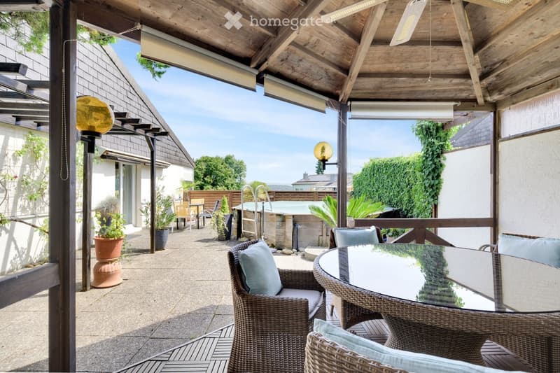 Covered outdoor patio with wooden beams, glass table, wicker chairs, cushions, and a small fountain.