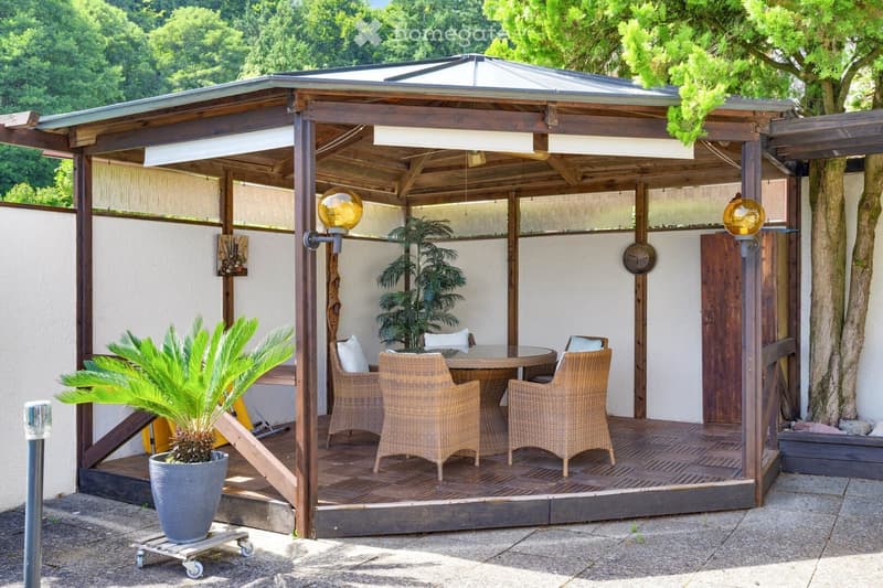 Wooden terrace with a covered roof, glass walls, and a dining set including a table and four chairs. Potted plants are placed around the area.