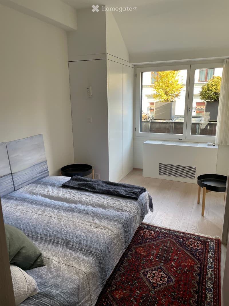 Cozy bedroom with a double bed, pillows, blanket, a small stool, and a patterned rug on light wooden flooring. Large windows offer a view of the outside, and built-in storage is visible on the wall.