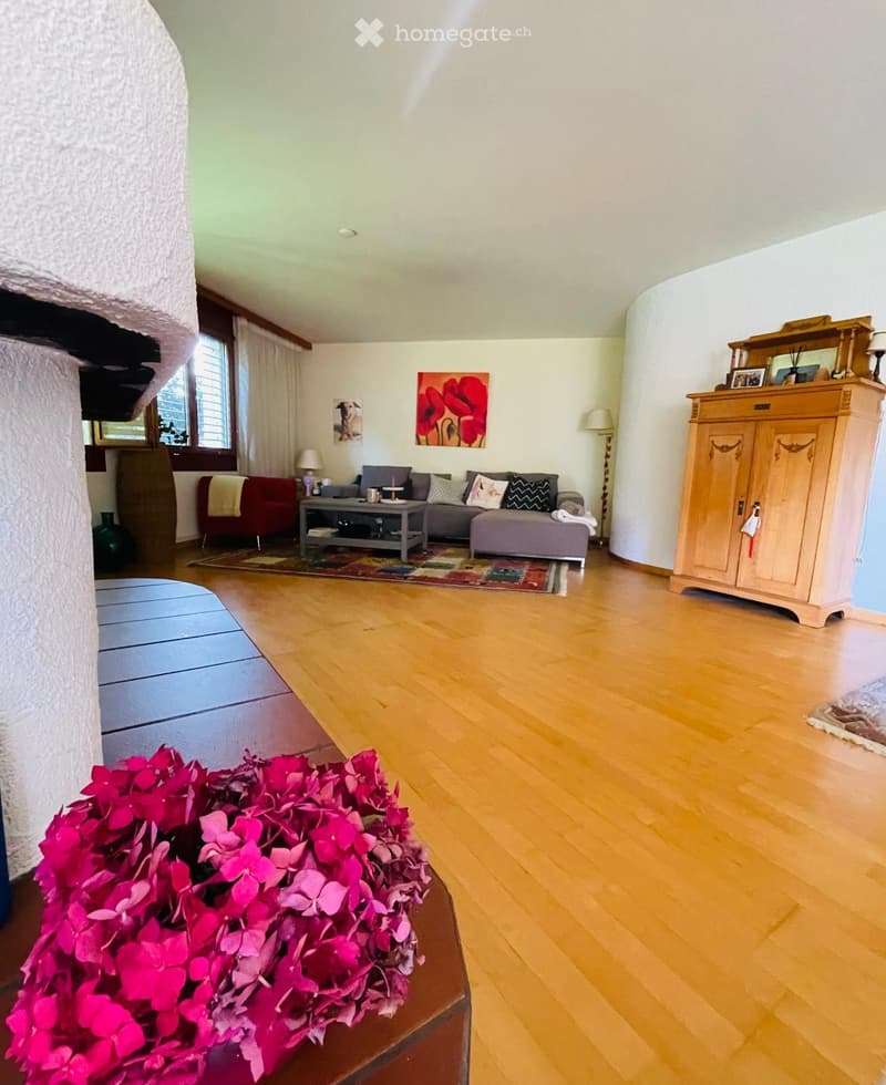 Bright living room with wooden flooring, a grey sofa, a red armchair, a coffee table, a wooden cabinet, and a floor lamp. A colorful rug is placed in front of the seating area.
