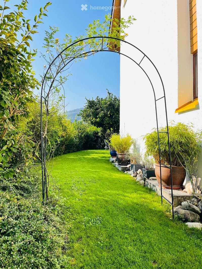 Well-maintained garden with a green lawn, potted plants, and a decorative metal archway leading to a view of the surrounding landscape.