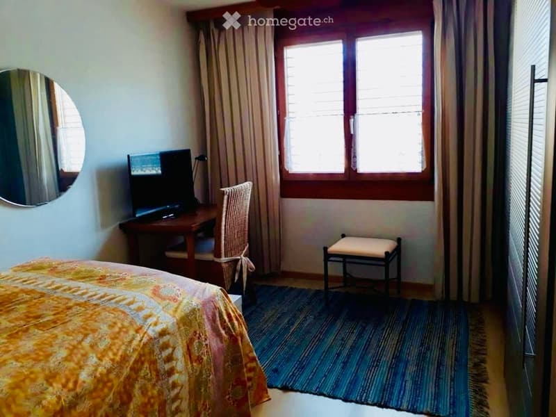Cozy bedroom with a bed, desk, chair, stool, mirror, and television. The room features a colorful bedspread, a patterned rug, and large windows with curtains.