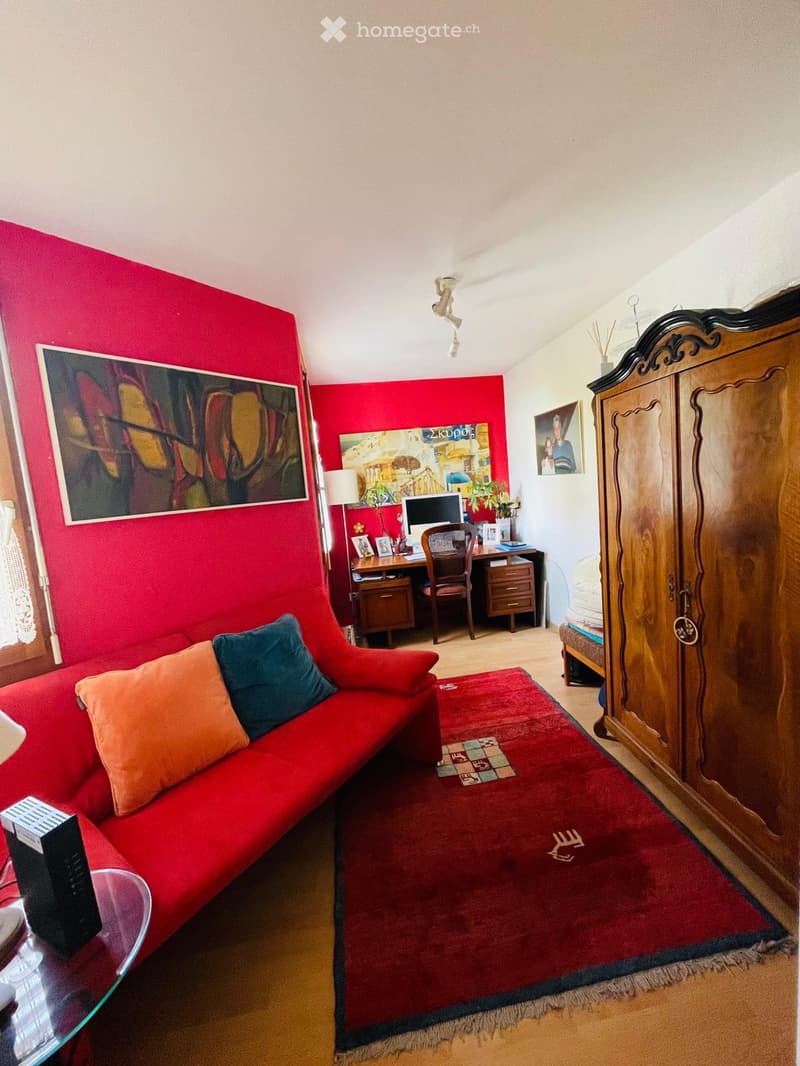 A cozy living room with a red sofa, colorful cushions, a wooden cabinet, a desk with a computer, and artwork on the walls.